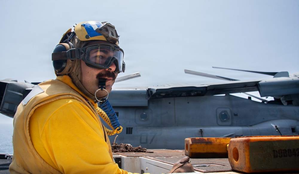 A naval deck crew member wearing a yellow jersey, flight deck helmet, ear protection, and safety goggles is positioned on what appears to be an amphibious assault ship or aircraft carrier flight deck. Behind him is a folded-rotor military tiltrotor aircraft, likely an MV-22 Osprey, with its rotors folded for deck storage. Orange chocks and chains are visible in the foreground, indicating aircraft tie-down operations.