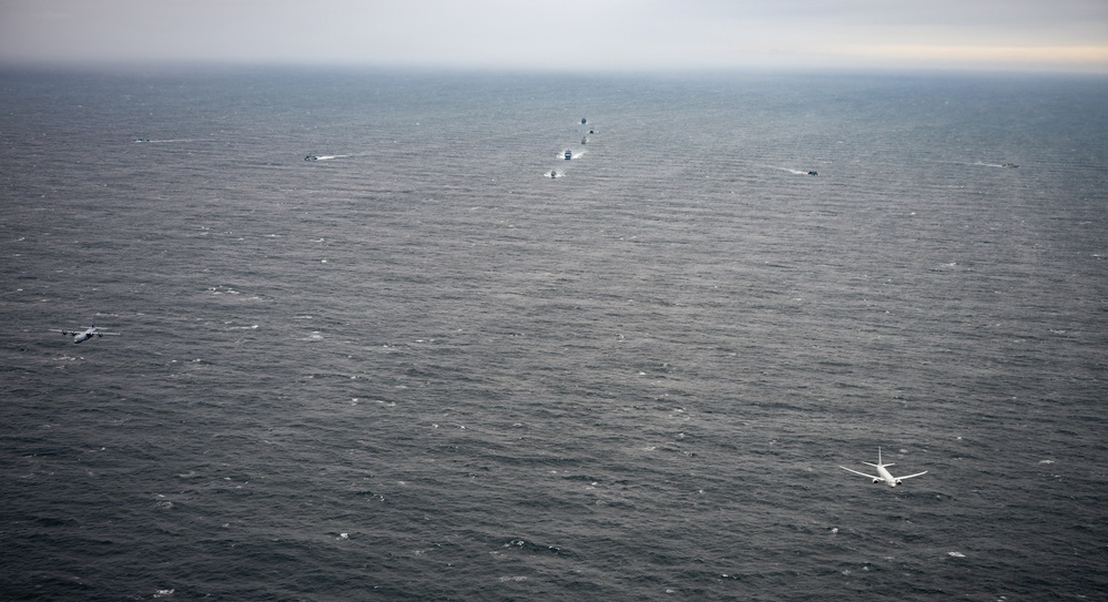 An aerial photograph taken over a large body of open ocean under overcast skies. Two military or patrol aircraft are flying low over the water, while a line of several vessels is visible in the distance forming a loose convoy or search formation. The scene suggests a coordinated maritime patrol or search-and-rescue operation.