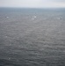 An aerial photograph taken over a large body of open ocean under overcast skies. Two military or patrol aircraft are flying low over the water, while a line of several vessels is visible in the distance forming a loose convoy or search formation. The scene suggests a coordinated maritime patrol or search-and-rescue operation.