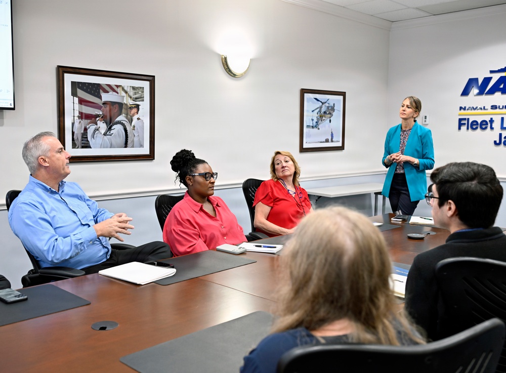 A professional meeting is taking place in a Naval Supply Fleet Logistics command conference room in Jacksonville. A woman in a teal blazer is standing and presenting to a group of five seated attendees around a wooden conference table. The setting is a formal government/military civilian workplace environment.
