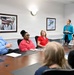 A professional meeting is taking place in a Naval Supply Fleet Logistics command conference room in Jacksonville. A woman in a teal blazer is standing and presenting to a group of five seated attendees around a wooden conference table. The setting is a formal government/military civilian workplace environment.