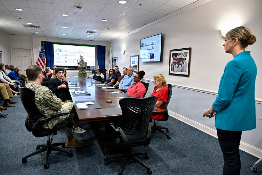 A formal briefing or meeting is taking place in a military conference room, with a mix of uniformed Navy personnel and civilian attendees seated around a large conference table. A woman in a teal blazer appears to be presenting or facilitating the meeting, while a uniformed service member stands near a projection screen at the far end of the room. An American flag and a blue military flag are visible in the background.