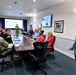 A formal briefing or meeting is taking place in a military conference room, with a mix of uniformed Navy personnel and civilian attendees seated around a large conference table. A woman in a teal blazer appears to be presenting or facilitating the meeting, while a uniformed service member stands near a projection screen at the far end of the room. An American flag and a blue military flag are visible in the background.