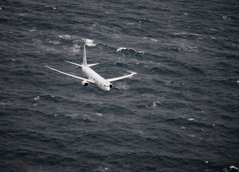 A white military or patrol aircraft is flying low over a rough, dark ocean. The aircraft appears to be a Boeing P-8 Poseidon or similar maritime patrol aircraft, photographed from above or at a similar altitude. The sea conditions appear turbulent with visible whitecaps and choppy waves.