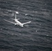A white military or patrol aircraft is flying low over a rough, dark ocean. The aircraft appears to be a Boeing P-8 Poseidon or similar maritime patrol aircraft, photographed from above or at a similar altitude. The sea conditions appear turbulent with visible whitecaps and choppy waves.