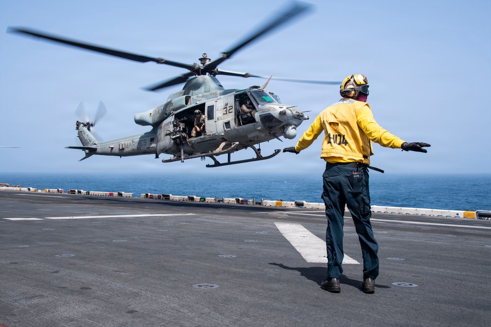 A U.S. Marines helicopter, marked with the number 32, is hovering just above the flight deck of a naval vessel while a flight deck crew member in a yellow jersey directs the aircraft. The scene takes place on what appears to be an amphibious assault ship or similar naval vessel, with open ocean visible in the background. The yellow-shirted handler is using hand signals to guide the helicopter during takeoff or landing operations.