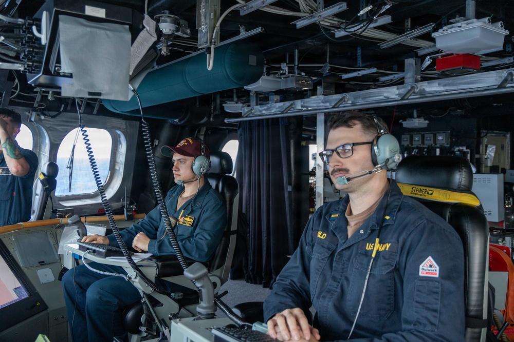 Two U.S. Navy personnel are stationed at control consoles aboard what appears to be the bridge or combat information center of a naval vessel. Both individuals are wearing communication headsets and Navy working uniforms (NWUs), with one wearing a maroon baseball cap. The environment is densely equipped with electronics, cables, monitors, and overhead equipment typical of a modern warship's operational spaces.