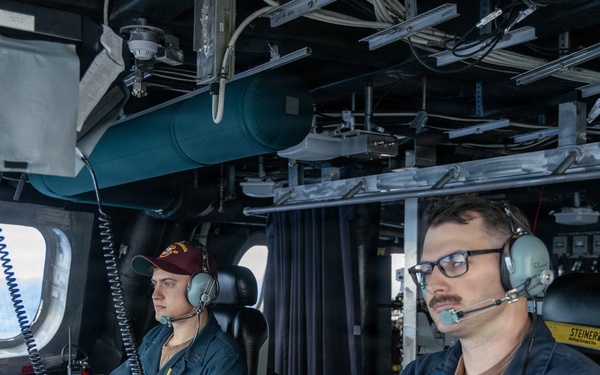 Two U.S. Navy personnel are stationed at control consoles aboard what appears to be the bridge or combat information center of a naval vessel. Both individuals are wearing communication headsets and Navy working uniforms (NWUs), with one wearing a maroon baseball cap. The environment is densely equipped with electronics, cables, monitors, and overhead equipment typical of a modern warship's operational spaces.