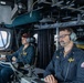 Two U.S. Navy personnel are stationed at control consoles aboard what appears to be the bridge or combat information center of a naval vessel. Both individuals are wearing communication headsets and Navy working uniforms (NWUs), with one wearing a maroon baseball cap. The environment is densely equipped with electronics, cables, monitors, and overhead equipment typical of a modern warship's operational spaces.
