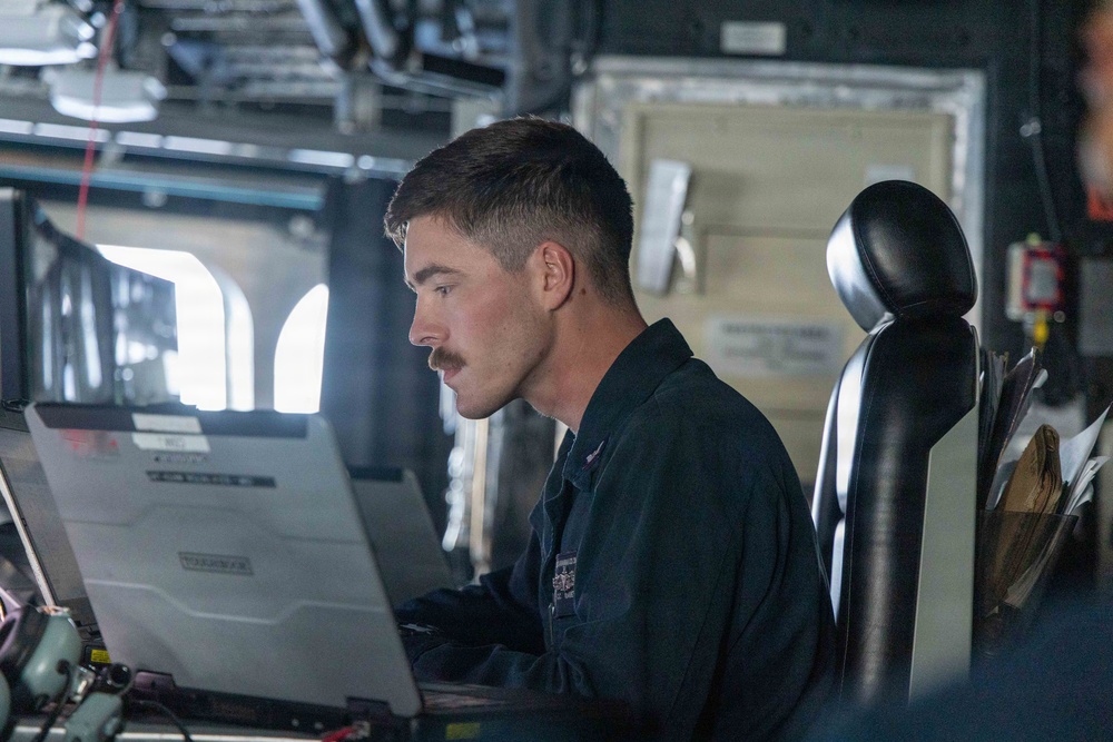 A young male military personnel member with a mustache is seated at a workstation aboard what appears to be a naval vessel, focused on a ruggedized laptop computer. He is wearing a dark navy coverall uniform with visible patches/insignia. The setting appears to be a ship's bridge or combat information center.