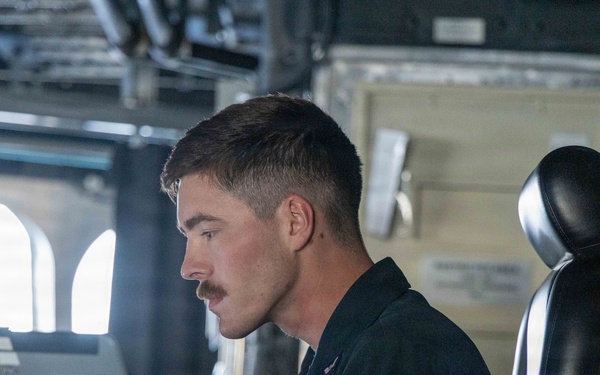 A young male military personnel member with a mustache is seated at a workstation aboard what appears to be a naval vessel, focused on a ruggedized laptop computer. He is wearing a dark navy coverall uniform with visible patches/insignia. The setting appears to be a ship's bridge or combat information center.