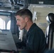 A young male military personnel member with a mustache is seated at a workstation aboard what appears to be a naval vessel, focused on a ruggedized laptop computer. He is wearing a dark navy coverall uniform with visible patches/insignia. The setting appears to be a ship's bridge or combat information center.