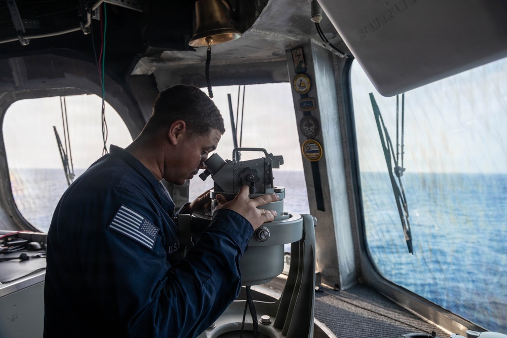 A U.S. Navy sailor is using a gyrocompass or alidade bearing instrument on the bridge of a naval vessel at sea. The sailor is wearing a navy blue uniform with a visible American flag patch and 'U.S.N.' lettering. The bridge environment shows open ocean visible through the windows, navigation equipment, and unit insignia patches on the bulkhead.