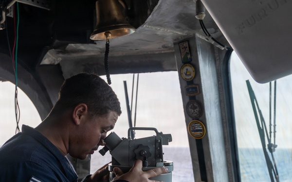 A U.S. Navy sailor is using a gyrocompass or alidade bearing instrument on the bridge of a naval vessel at sea. The sailor is wearing a navy blue uniform with a visible American flag patch and 'U.S.N.' lettering. The bridge environment shows open ocean visible through the windows, navigation equipment, and unit insignia patches on the bulkhead.