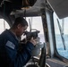 A U.S. Navy sailor is using a gyrocompass or alidade bearing instrument on the bridge of a naval vessel at sea. The sailor is wearing a navy blue uniform with a visible American flag patch and 'U.S.N.' lettering. The bridge environment shows open ocean visible through the windows, navigation equipment, and unit insignia patches on the bulkhead.