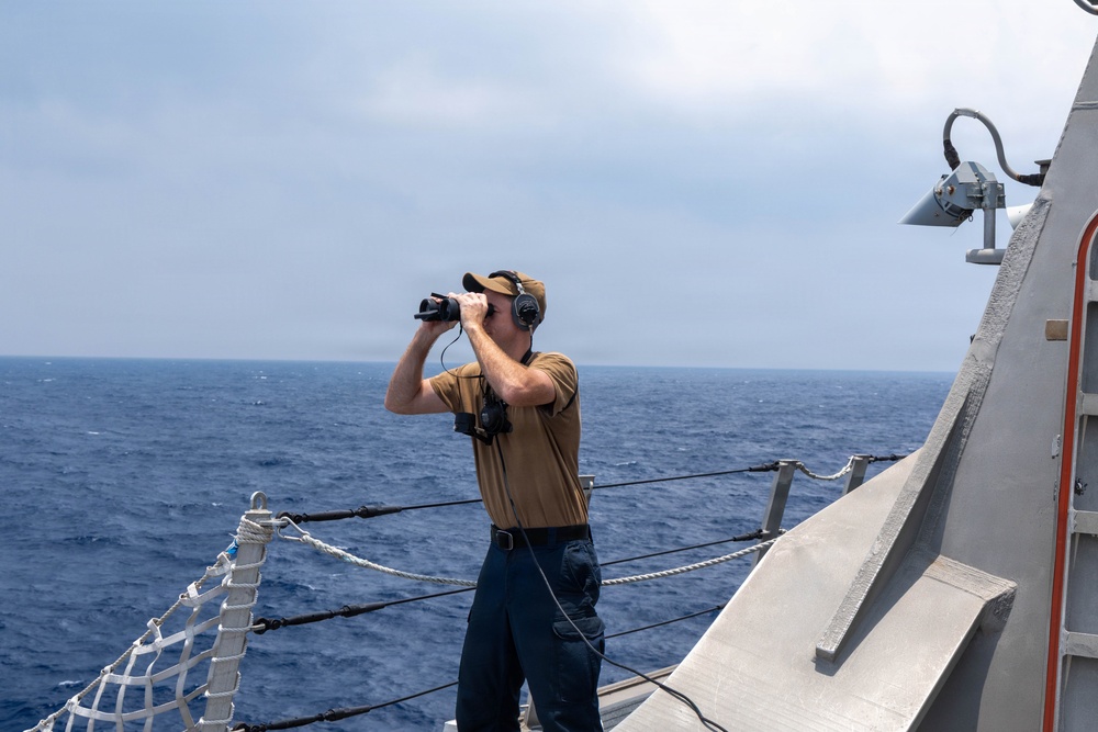 A naval personnel member stands on the deck of a military vessel, using binoculars to scan the open ocean. The individual is equipped with communication headphones and a radio device, suggesting an active watch or lookout duty. The vessel appears to be a modern warship operating in open blue water under hazy conditions.
