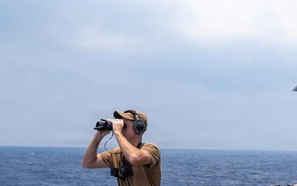 A naval personnel member stands on the deck of a military vessel, using binoculars to scan the open ocean. The individual is equipped with communication headphones and a radio device, suggesting an active watch or lookout duty. The vessel appears to be a modern warship operating in open blue water under hazy conditions.