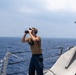 A naval personnel member stands on the deck of a military vessel, using binoculars to scan the open ocean. The individual is equipped with communication headphones and a radio device, suggesting an active watch or lookout duty. The vessel appears to be a modern warship operating in open blue water under hazy conditions.