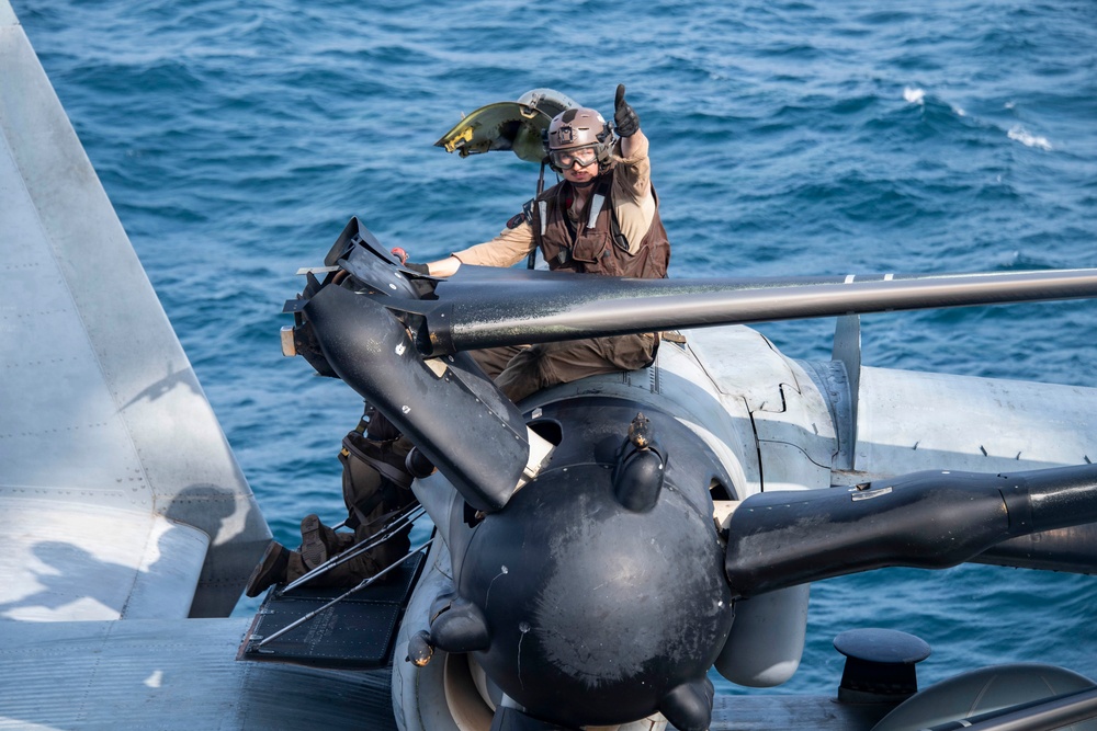 A military aviation crew member in flight gear is positioned on or near a naval aircraft on what appears to be a ship deck over open ocean. The individual is giving a thumbs-up gesture, likely signaling readiness or an all-clear status. The scene depicts standard carrier or naval vessel flight deck operations.