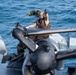 A military aviation crew member in flight gear is positioned on or near a naval aircraft on what appears to be a ship deck over open ocean. The individual is giving a thumbs-up gesture, likely signaling readiness or an all-clear status. The scene depicts standard carrier or naval vessel flight deck operations.