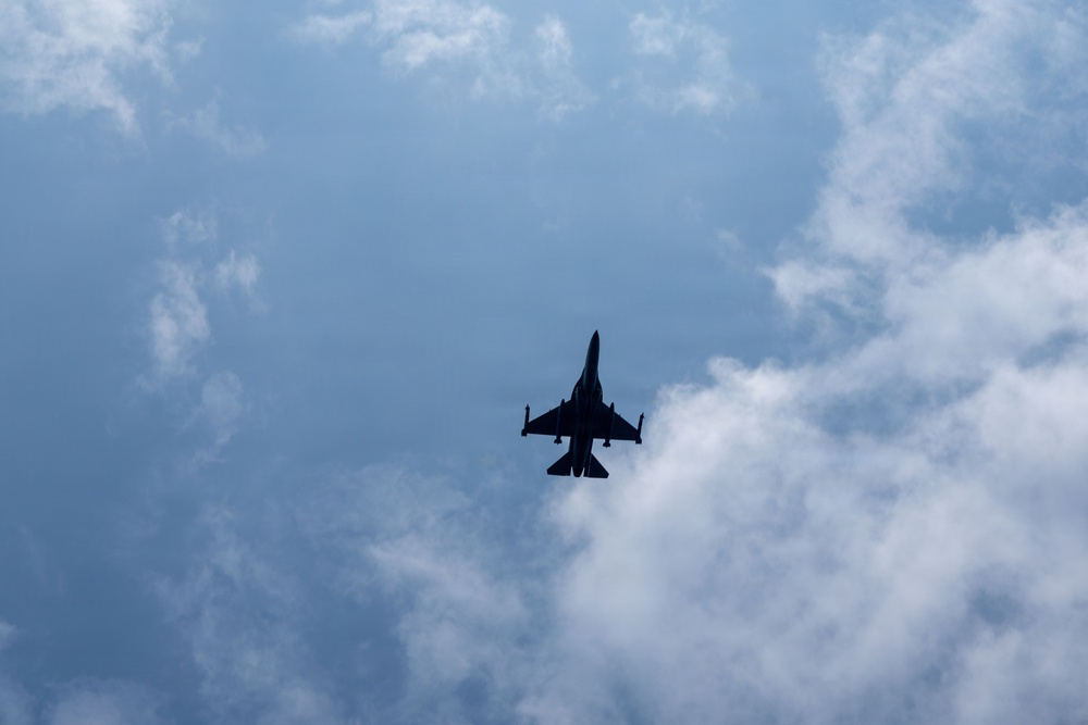 A military jet fighter is captured in silhouette from a ground-level upward perspective against a partly cloudy blue sky. The aircraft appears to be in level flight, viewed from directly below, revealing its underside profile. The backlighting creates a stark dark silhouette against the bright sky.