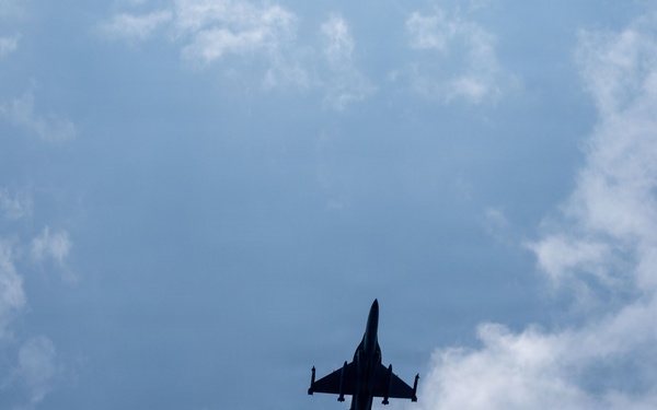 A military jet fighter is captured in silhouette from a ground-level upward perspective against a partly cloudy blue sky. The aircraft appears to be in level flight, viewed from directly below, revealing its underside profile. The backlighting creates a stark dark silhouette against the bright sky.