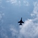 A military jet fighter is captured in silhouette from a ground-level upward perspective against a partly cloudy blue sky. The aircraft appears to be in level flight, viewed from directly below, revealing its underside profile. The backlighting creates a stark dark silhouette against the bright sky.