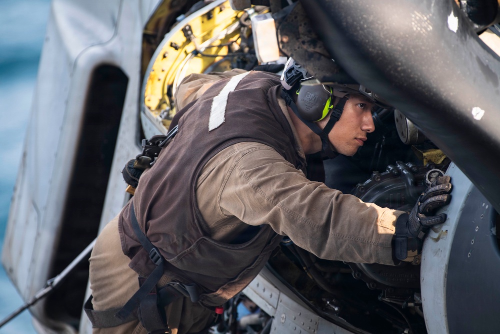 A military or aviation technician is performing maintenance work on what appears to be a helicopter, leaning into an open access panel or engine compartment. The individual is wearing hearing protection, tactical gloves, and a brown flight suit or work uniform with a dark vest. The background suggests an outdoor maritime or flight deck environment.