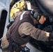 A military or aviation technician is performing maintenance work on what appears to be a helicopter, leaning into an open access panel or engine compartment. The individual is wearing hearing protection, tactical gloves, and a brown flight suit or work uniform with a dark vest. The background suggests an outdoor maritime or flight deck environment.