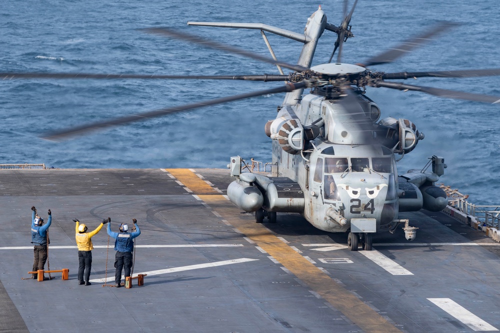 A large military helicopter marked with the number '24' is on the flight deck of a naval vessel, with rotor blades spinning. Three flight deck crew members in colored jerseys are signaling with raised arms, likely directing the helicopter for takeoff or landing operations. The scene takes place at sea with open ocean visible in the background.