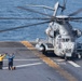 A large military helicopter marked with the number '24' is on the flight deck of a naval vessel, with rotor blades spinning. Three flight deck crew members in colored jerseys are signaling with raised arms, likely directing the helicopter for takeoff or landing operations. The scene takes place at sea with open ocean visible in the background.