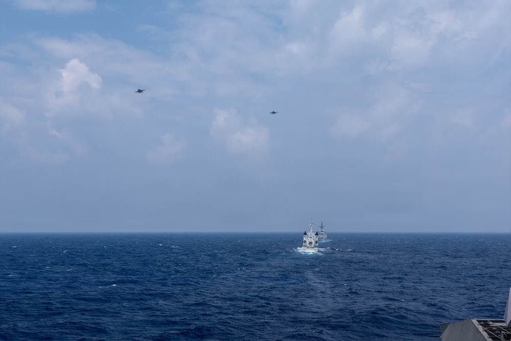 A naval scene captured from the deck of a warship showing multiple vessels underway on open ocean. Two military aircraft are visible flying overhead in the distance above the formation of ships. The scene appears to depict a coordinated naval exercise or transit operation.