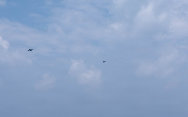 A naval scene captured from the deck of a warship showing multiple vessels underway on open ocean. Two military aircraft are visible flying overhead in the distance above the formation of ships. The scene appears to depict a coordinated naval exercise or transit operation.