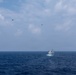 A naval scene captured from the deck of a warship showing multiple vessels underway on open ocean. Two military aircraft are visible flying overhead in the distance above the formation of ships. The scene appears to depict a coordinated naval exercise or transit operation.