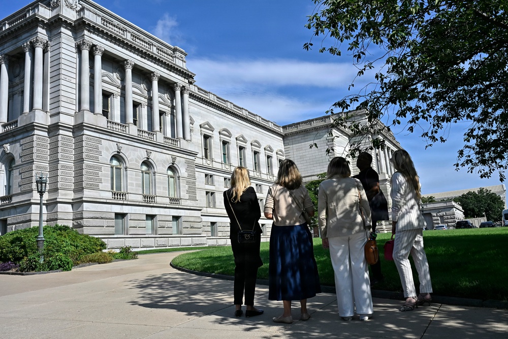 A group of five people stand with their backs to the camera, looking toward a large neoclassical government building with columns, arched windows, and ornate stone detailing. The scene appears to be an outdoor tour or sightseeing visit on a clear, sunny day. The building's architecture and surrounding landscaping suggest a prominent civic or governmental institution.