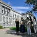 A group of five people stand with their backs to the camera, looking toward a large neoclassical government building with columns, arched windows, and ornate stone detailing. The scene appears to be an outdoor tour or sightseeing visit on a clear, sunny day. The building's architecture and surrounding landscaping suggest a prominent civic or governmental institution.