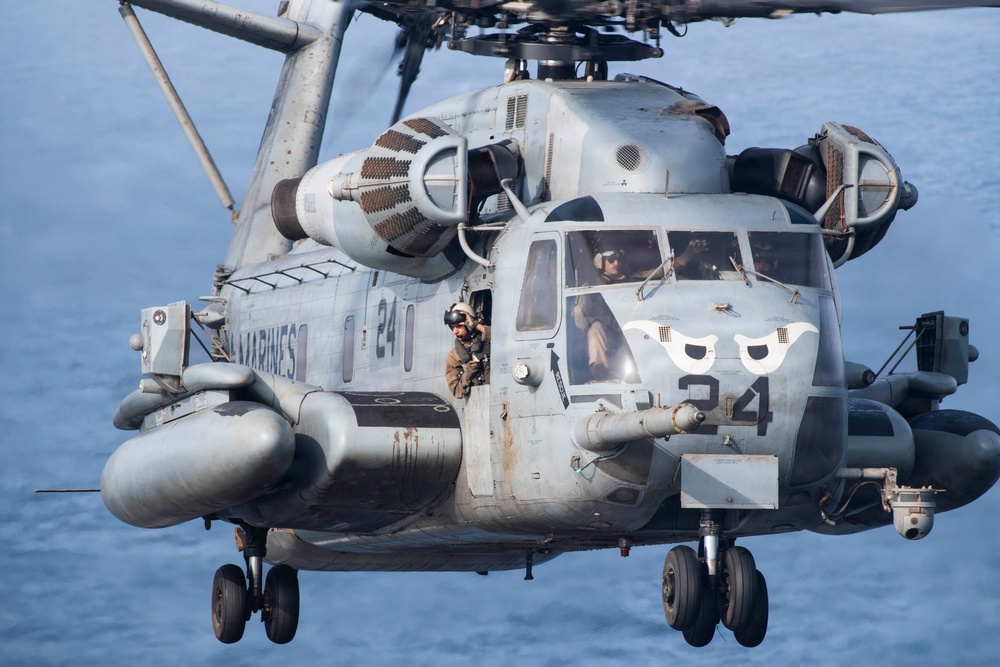 A U.S. Marines heavy-lift helicopter, identified as aircraft number 24, is captured in close-up flight over open water. The helicopter displays a distinctive owl-face nose art and bears the 'MARINES' marking on its fuselage. A crew member is visible leaning out of an open side door while pilots are seen through the cockpit windows.