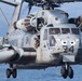 A U.S. Marines heavy-lift helicopter, identified as aircraft number 24, is captured in close-up flight over open water. The helicopter displays a distinctive owl-face nose art and bears the 'MARINES' marking on its fuselage. A crew member is visible leaning out of an open side door while pilots are seen through the cockpit windows.