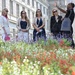 A group of four women listens attentively to a man who appears to be a tour guide, gesturing upward toward a large neoclassical building. The scene takes place in front of a colorful garden bed filled with red, white, and purple flowers. The man is carrying a tote bag labeled 'Capital Jewish Museum,' suggesting this is an organized tour.