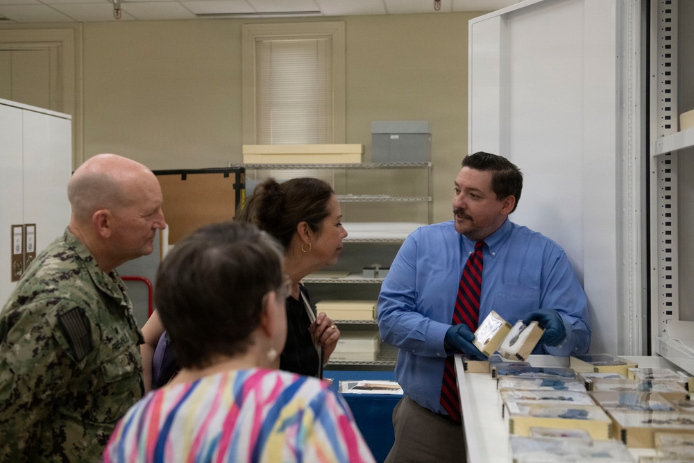 A man in a blue shirt and striped tie, wearing blue nitrile gloves, appears to be presenting or explaining archival materials to a small group that includes a U.S. Navy officer in camouflage uniform and two civilian women. The setting appears to be an archival storage or processing room with shelving units and flat storage boxes visible. The man is holding what appears to be a small archival box or item from a tray of similar materials on the table.
