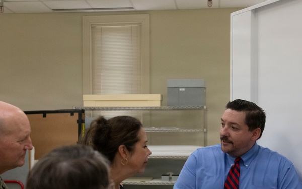 A man in a blue shirt and striped tie, wearing blue nitrile gloves, appears to be presenting or explaining archival materials to a small group that includes a U.S. Navy officer in camouflage uniform and two civilian women. The setting appears to be an archival storage or processing room with shelving units and flat storage boxes visible. The man is holding what appears to be a small archival box or item from a tray of similar materials on the table.