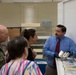 A man in a blue shirt and striped tie, wearing blue nitrile gloves, appears to be presenting or explaining archival materials to a small group that includes a U.S. Navy officer in camouflage uniform and two civilian women. The setting appears to be an archival storage or processing room with shelving units and flat storage boxes visible. The man is holding what appears to be a small archival box or item from a tray of similar materials on the table.
