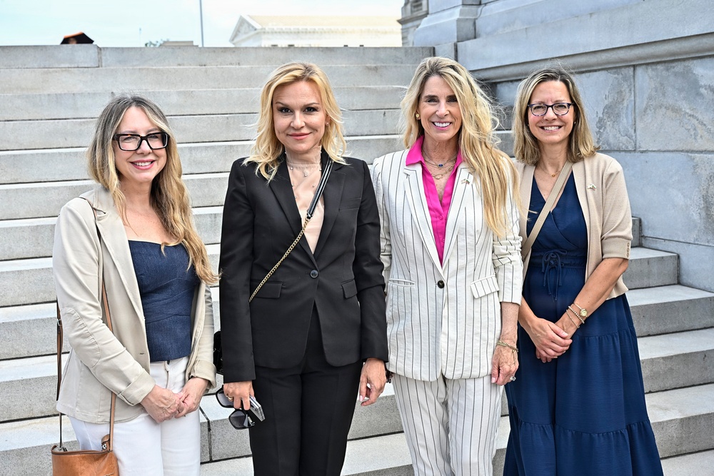 Four women are posing together on the steps of what appears to be a government or institutional building, likely in Washington D.C. based on the neoclassical architecture visible in the background. They are dressed in professional business attire and appear to be at a formal or official event. All four are smiling and facing the camera in a group photo.