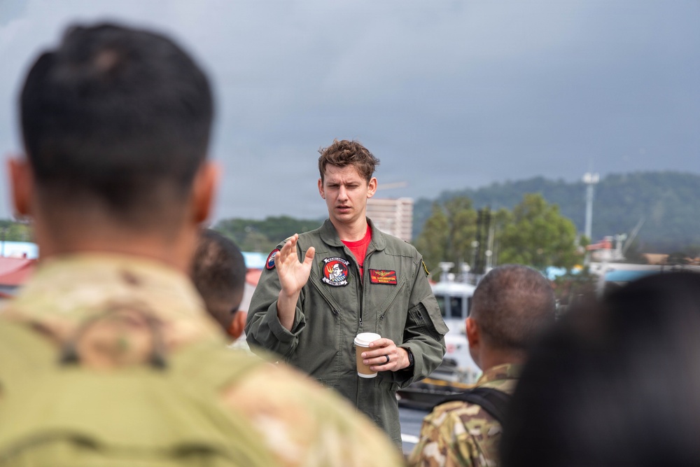 A military aviator in an olive green flight suit is addressing a small group of soldiers in camouflage uniforms outdoors. He is holding a disposable coffee cup and gesturing with one hand, suggesting he is briefing or instructing the group. The setting appears to be a port or waterfront area with overcast skies and urban infrastructure visible in the background.
