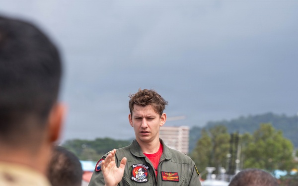 A military aviator in an olive green flight suit is addressing a small group of soldiers in camouflage uniforms outdoors. He is holding a disposable coffee cup and gesturing with one hand, suggesting he is briefing or instructing the group. The setting appears to be a port or waterfront area with overcast skies and urban infrastructure visible in the background.
