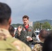 A military aviator in an olive green flight suit is addressing a small group of soldiers in camouflage uniforms outdoors. He is holding a disposable coffee cup and gesturing with one hand, suggesting he is briefing or instructing the group. The setting appears to be a port or waterfront area with overcast skies and urban infrastructure visible in the background.