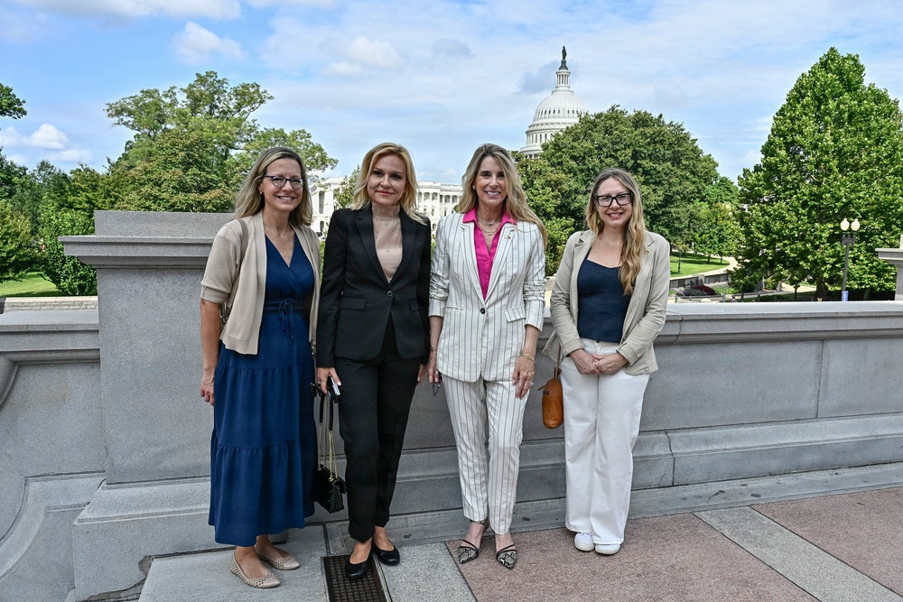 Four women are posing together for a photo outdoors on what appears to be a terrace or plaza near the United States Capitol building in Washington, D.C. The group is dressed in professional business attire, suggesting an official or formal visit. The setting, lush greenery, and clear sky indicate a warm-weather daytime event.