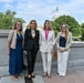 Four women are posing together for a photo outdoors on what appears to be a terrace or plaza near the United States Capitol building in Washington, D.C. The group is dressed in professional business attire, suggesting an official or formal visit. The setting, lush greenery, and clear sky indicate a warm-weather daytime event.