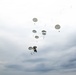 The image shows a large-scale military airborne operation with numerous parachutes deployed across an overcast sky. Multiple round, olive/gray-colored parachutes are visible at various altitudes, suggesting a mass tactical jump or airdrop in progress. At least one aircraft is faintly visible in the lower left background, likely the delivery platform.