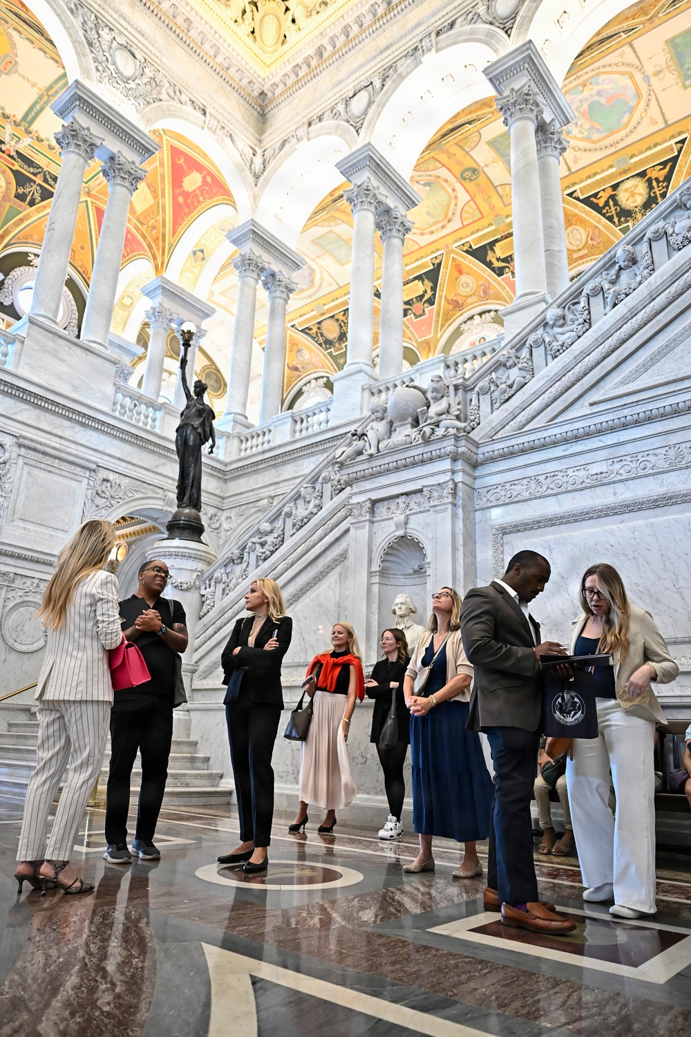 A group of approximately ten visitors are gathered in the ornate Great Hall of the Library of Congress Thomas Jefferson Building in Washington, D.C. The space features elaborate marble staircases, Corinthian columns, richly painted vaulted ceilings in gold, red, and green, and a bronze statue of a torch-bearing figure. The visitors appear to be on a guided tour, with some looking upward at the architecture and one person reviewing materials on a tablet.